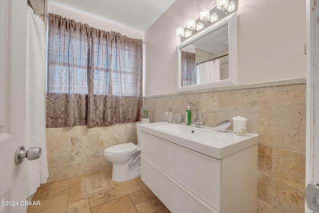 full bathroom with tile walls, stone tile floors, toilet, wainscoting, and vanity