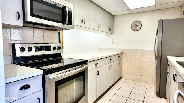 kitchen with tile walls, stainless steel appliances, white cabinetry, light tile patterned floors, and a drop ceiling