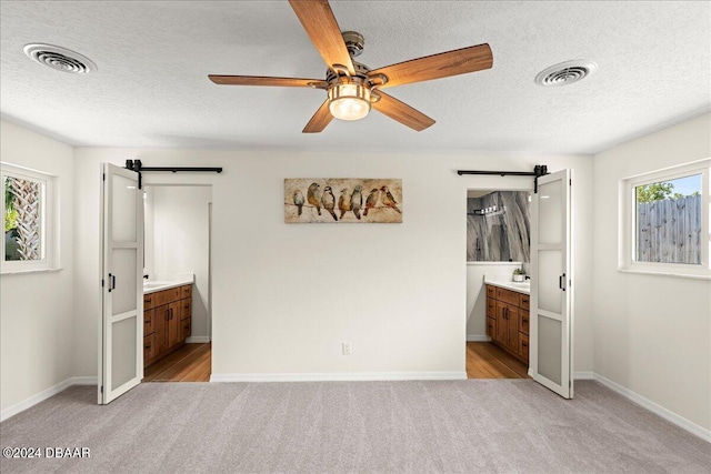 unfurnished bedroom featuring a barn door, light colored carpet, ceiling fan, and ensuite bath