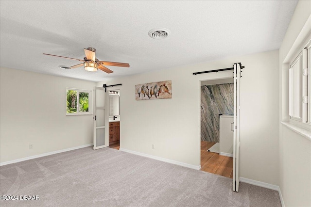 unfurnished bedroom with light colored carpet, a textured ceiling, a barn door, connected bathroom, and ceiling fan