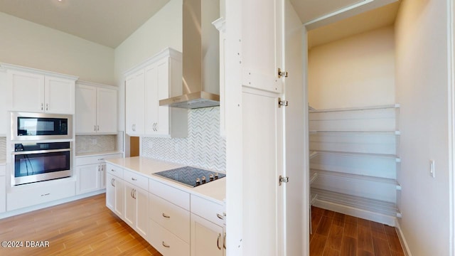 kitchen featuring light wood finished floors, built in microwave, stainless steel oven, wall chimney exhaust hood, and black electric cooktop