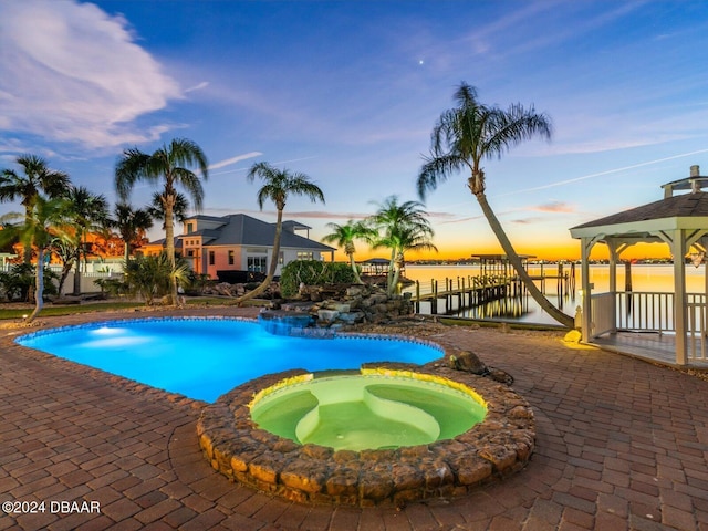 pool at dusk featuring an in ground hot tub, a water view, and a patio