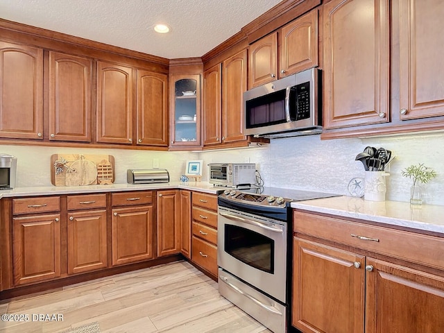 kitchen with brown cabinets, a textured ceiling, stainless steel appliances, light wood finished floors, and glass insert cabinets