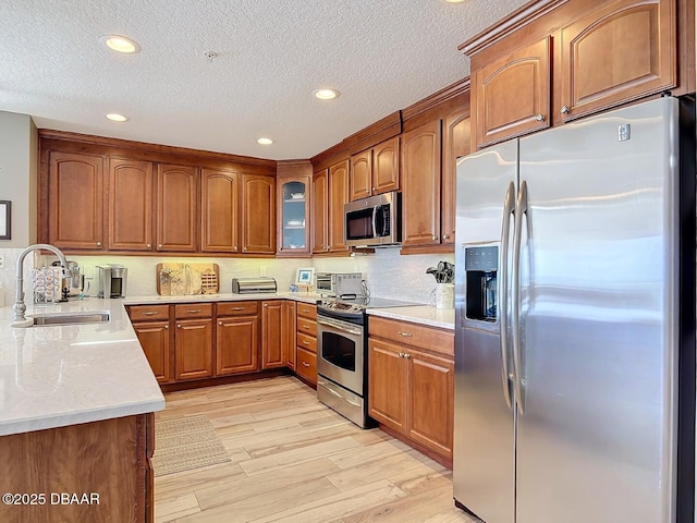 kitchen with brown cabinetry, light wood-style flooring, a sink, glass insert cabinets, and appliances with stainless steel finishes