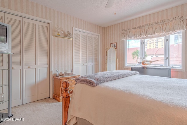 bedroom featuring wallpapered walls, two closets, ceiling fan, light colored carpet, and a textured ceiling