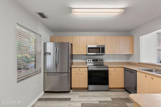 kitchen with light brown cabinetry, a textured ceiling, stainless steel appliances, sink, and light hardwood / wood-style floors