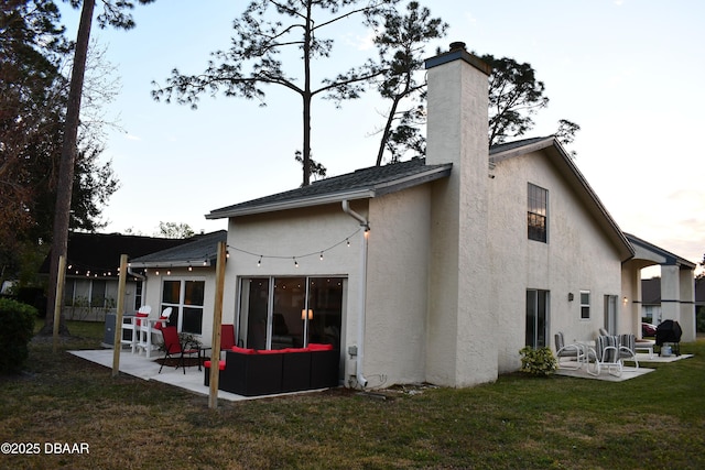 back of house with stucco siding, a lawn, a patio, roof with shingles, and a chimney