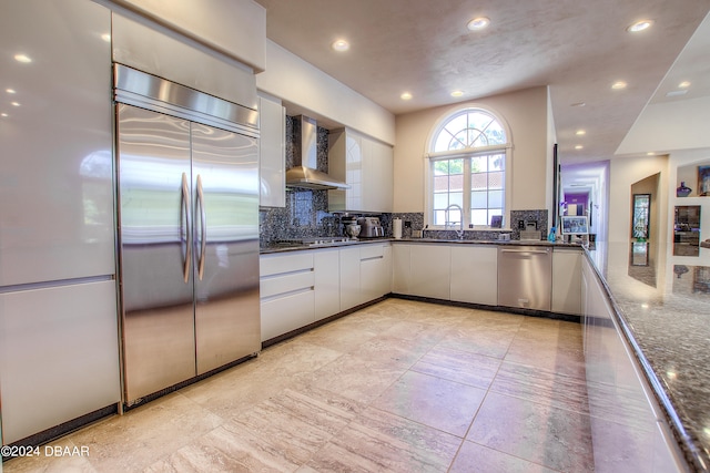 kitchen with stainless steel appliances, white cabinetry, wall chimney range hood, decorative backsplash, and dark stone countertops