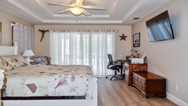 bedroom with a raised ceiling, multiple windows, a desk, and light wood-style flooring