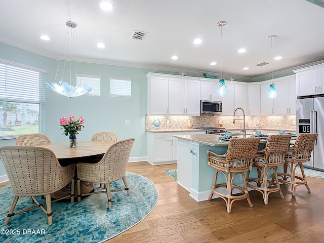 kitchen with light wood-type flooring, stainless steel appliances, decorative light fixtures, white cabinets, and an island with sink