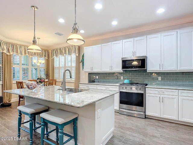 kitchen with stainless steel appliances, a sink, white cabinetry, hanging light fixtures, and a center island with sink