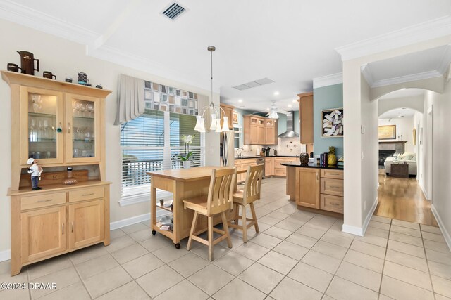 kitchen featuring crown molding, decorative light fixtures, light tile patterned floors, light brown cabinetry, and wall chimney exhaust hood