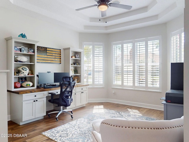 office space featuring ceiling fan, built in desk, a raised ceiling, baseboards, and light wood finished floors