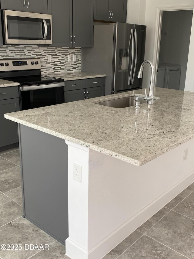 kitchen with appliances with stainless steel finishes, gray cabinetry, sink, light stone counters, and a center island with sink