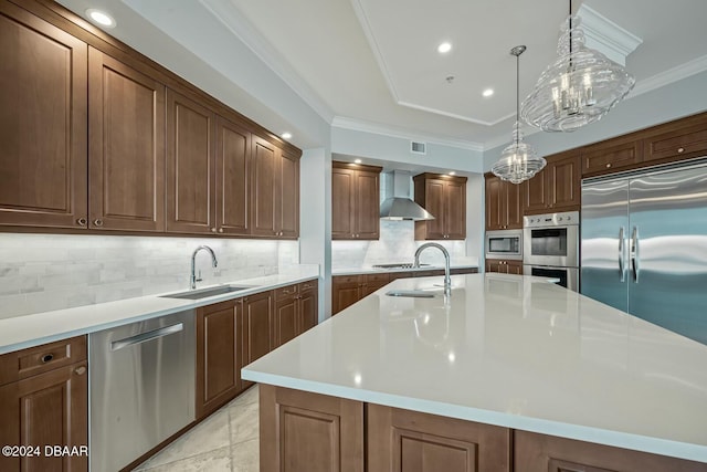 kitchen with wall chimney range hood, backsplash, built in appliances, an island with sink, and pendant lighting