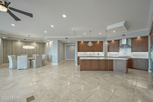 kitchen with pendant lighting, wall chimney range hood, crown molding, sink, and an island with sink