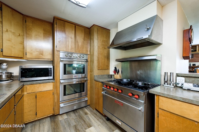 kitchen with a warming drawer, stainless steel appliances, brown cabinetry, wall chimney range hood, and light wood-type flooring