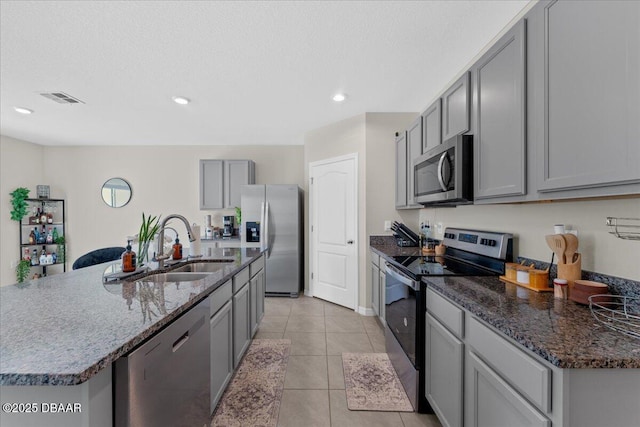 kitchen featuring light tile patterned flooring, gray cabinetry, stainless steel appliances, dark stone countertops, and an island with sink