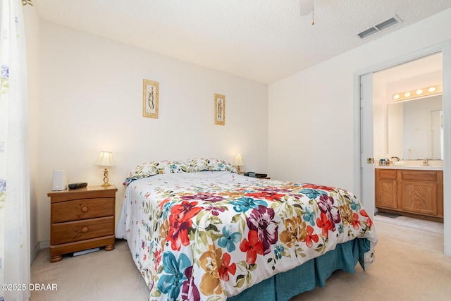 bedroom featuring visible vents, light colored carpet, a textured ceiling, ensuite bath, and a ceiling fan