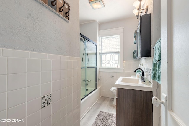 full bath with toilet, vanity, wood finished floors, shower / bath combination with glass door, and tile walls