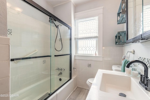 bathroom featuring wood finished floors, enclosed tub / shower combo, a sink, tile walls, and toilet