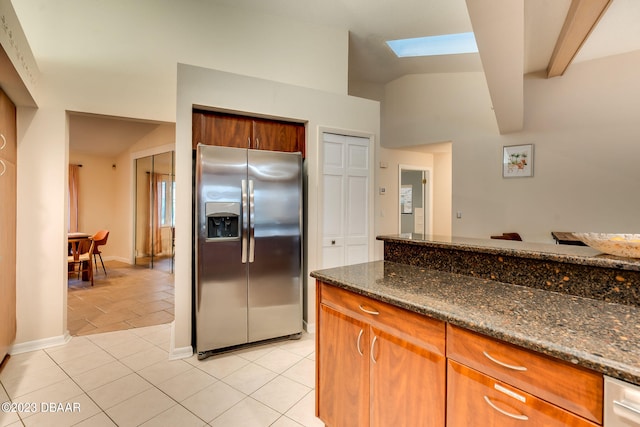 kitchen with stainless steel refrigerator with ice dispenser, dark stone countertops, light tile patterned floors, and lofted ceiling with skylight