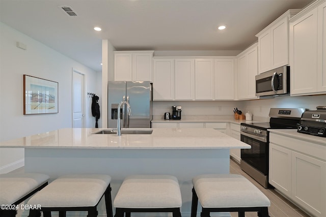 kitchen featuring light stone counters, appliances with stainless steel finishes, sink, white cabinets, and a kitchen island with sink