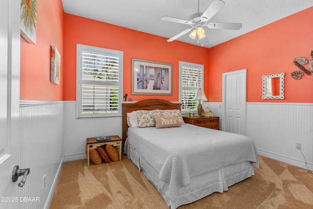 carpeted bedroom with a wainscoted wall, multiple windows, a textured ceiling, and a ceiling fan