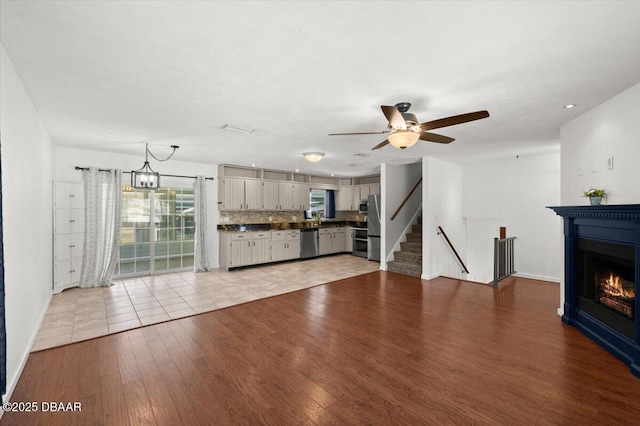 unfurnished living room with light wood-type flooring, ceiling fan with notable chandelier, a glass covered fireplace, stairway, and baseboards