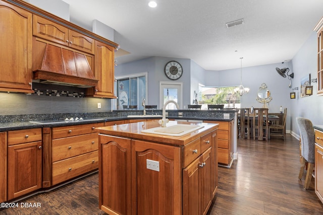 kitchen with dark hardwood / wood-style flooring, custom range hood, cooktop, a kitchen island with sink, and hanging light fixtures