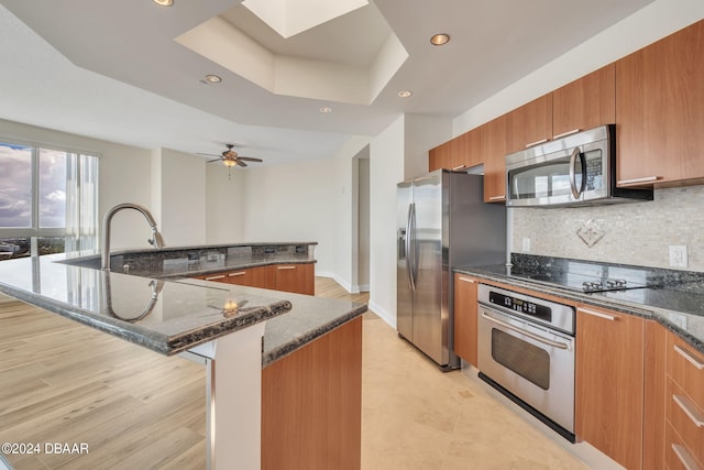 kitchen featuring a large island with sink, appliances with stainless steel finishes, tasteful backsplash, dark stone countertops, and a tray ceiling