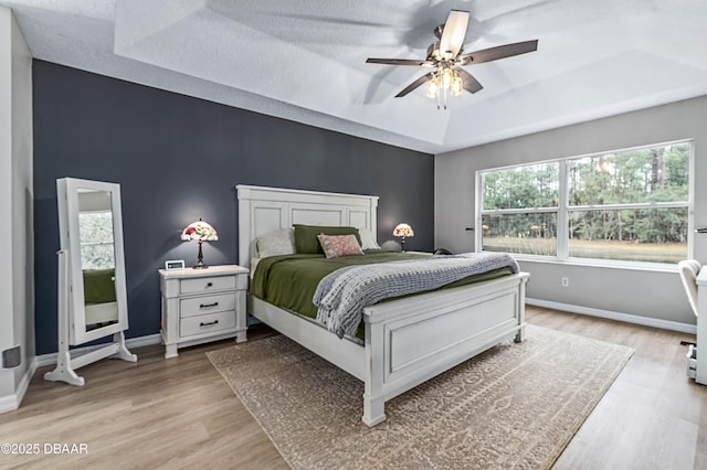 bedroom with light wood finished floors, baseboards, a raised ceiling, a ceiling fan, and an accent wall