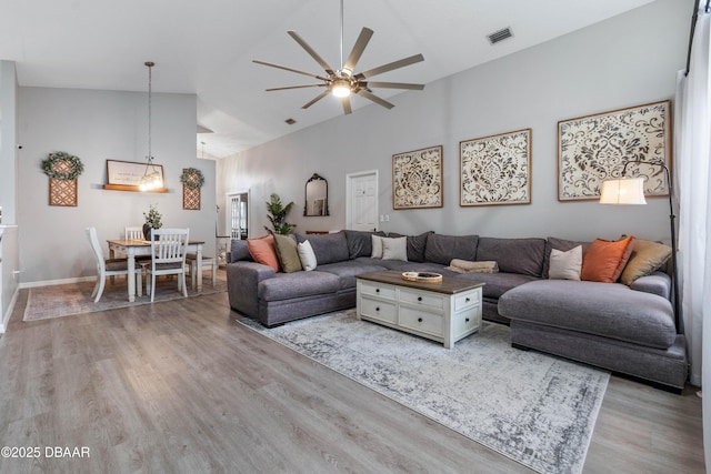 living area with visible vents, light wood-style floors, a ceiling fan, high vaulted ceiling, and baseboards