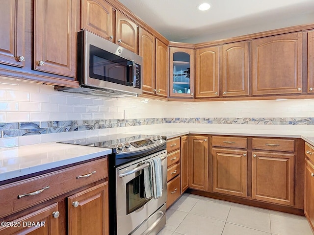 kitchen with glass insert cabinets, light tile patterned floors, appliances with stainless steel finishes, and brown cabinetry