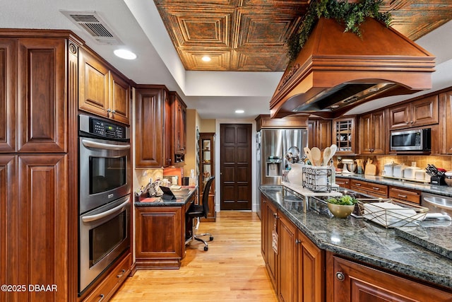 kitchen featuring custom exhaust hood, stainless steel appliances, built in desk, dark stone countertops, and light hardwood / wood-style floors