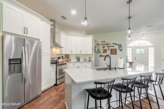 kitchen featuring appliances with stainless steel finishes, hanging light fixtures, an island with sink, white cabinets, and wall chimney range hood