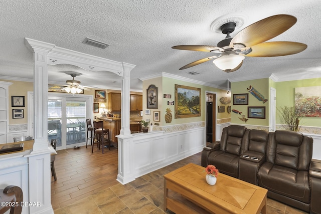 living area with visible vents, ceiling fan, a wainscoted wall, decorative columns, and a textured ceiling