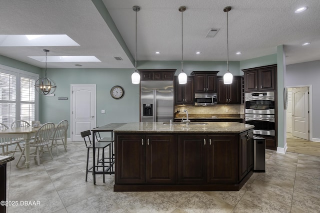 kitchen with light stone counters, dark brown cabinets, appliances with stainless steel finishes, pendant lighting, and a kitchen island with sink