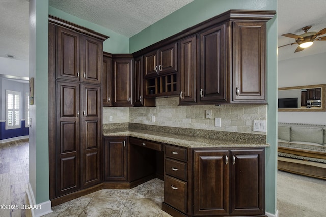 kitchen featuring dark brown cabinetry, backsplash, built in desk, and light stone countertops