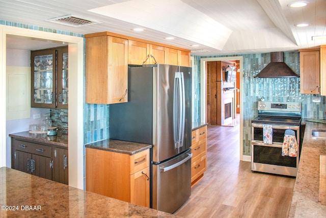 kitchen with stainless steel appliances, wall chimney range hood, tasteful backsplash, dark stone counters, and light wood-type flooring