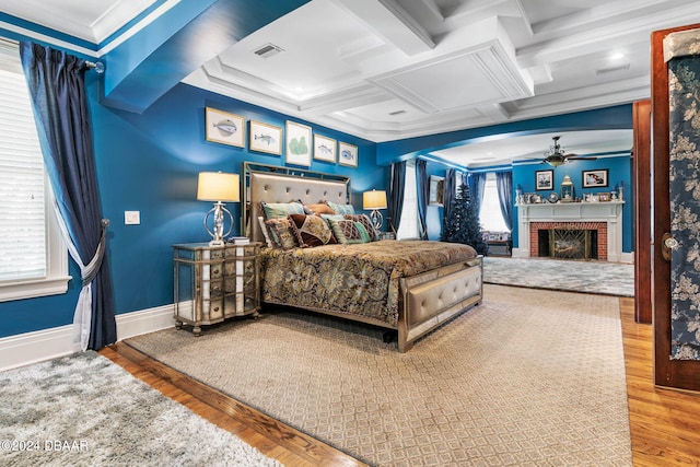 bedroom featuring wood-type flooring, crown molding, coffered ceiling, a brick fireplace, and beamed ceiling