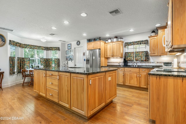 kitchen featuring stainless steel fridge with ice dispenser, a center island with sink, light hardwood / wood-style flooring, and dark stone countertops