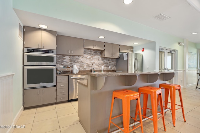 kitchen featuring a kitchen breakfast bar, gray cabinets, light tile patterned flooring, backsplash, and appliances with stainless steel finishes