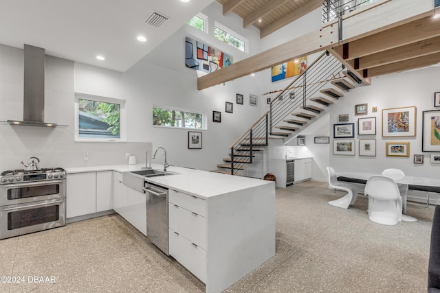 kitchen with a towering ceiling, stainless steel appliances, sink, wall chimney range hood, and beam ceiling