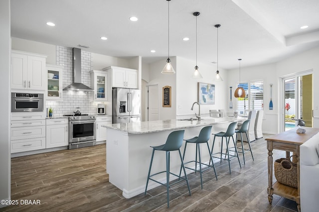 kitchen featuring white cabinetry, stainless steel appliances, wall chimney exhaust hood, decorative light fixtures, and a large island with sink