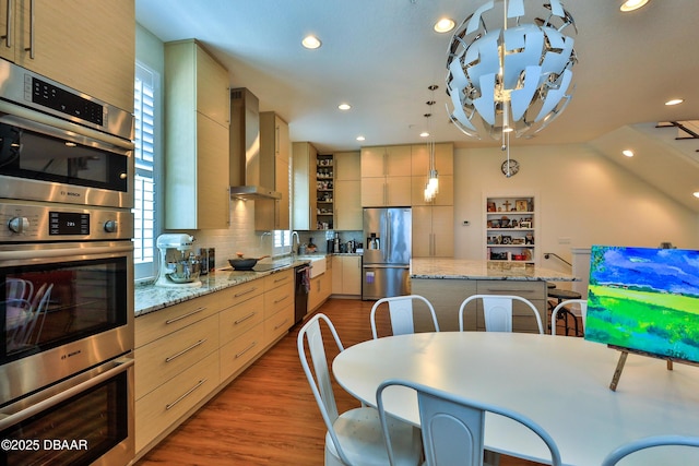 kitchen featuring light brown cabinetry, light stone counters, appliances with stainless steel finishes, pendant lighting, and wall chimney range hood