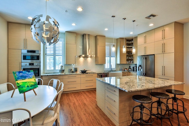 kitchen with wall chimney exhaust hood, light brown cabinetry, a kitchen island, pendant lighting, and stainless steel appliances