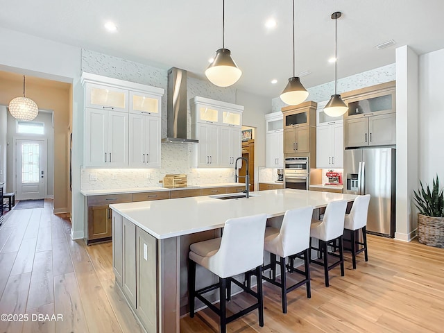 kitchen with light wood finished floors, stainless steel appliances, a sink, wall chimney range hood, and a large island with sink