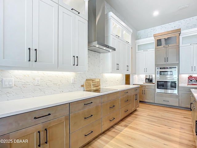 kitchen featuring stainless steel double oven, black electric cooktop, light countertops, wall chimney range hood, and light wood finished floors