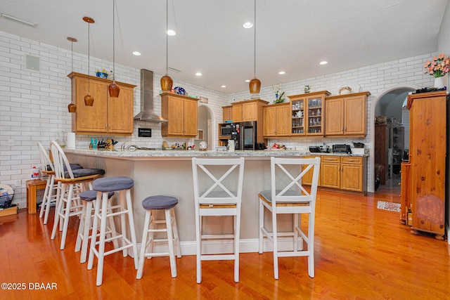 kitchen with brick wall, light wood-type flooring, hanging light fixtures, and wall chimney range hood